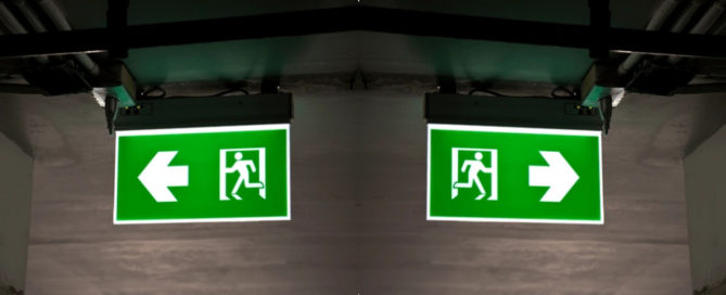 Señales de salida de emergencia iluminadas con flechas en ambos sentidos en un pasillo subterráneo